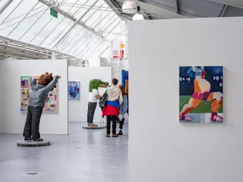 An art gallery with white walls displays colorful paintings and sculptures, including a figure with raised arms and another with a green textured head. Several visitors stand in the center viewing the artwork under a glass-paneled roof.