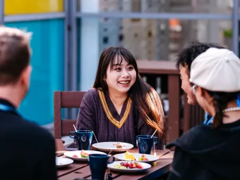 Students sitting outside at a table chatting. There are plates of food and mugs on the table.