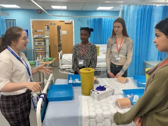 A group of students standing around a hospital bed, listening to a demonstrator.