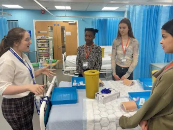 A group of students standing around a hospital bed, listening to a demonstrator.