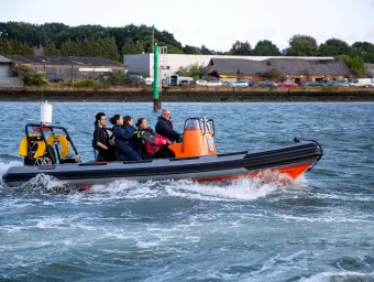 Students wearing life jackets ride a small orange RIB boat across the water during a summer school activity.
