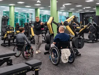 Two wheelchair users get advice from a personal trainer on how to use adaptive equipment in the gym. Other gym goers use benches and treadmills in the background