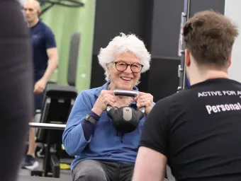 An elderly lady smiles at her instructor while lifting a kettle bell to her chest