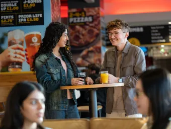 Two students chat over drinks in the Student Union. They're standing slightly to the side of each other, laughing together.