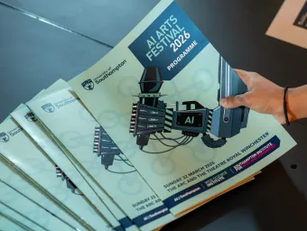 A hand picking up a programme for the University of Southampton AI Arts Festival 2026, from a stack of programmes on a table.