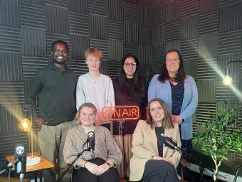 A group of podcast presenters in the studio. There are 2 presenters sitting and 4 presenters standing behind them.
