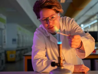 A foundation student in white lab coat and safety goggles lights a Bunsen burner in a lab