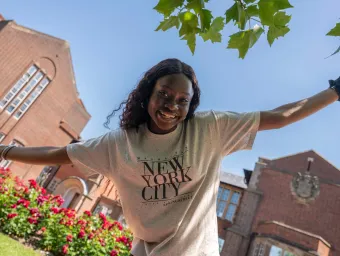 An international student poses in front of a university building, her arms outstretched, happy and welcoming, with a beaming smile. She's framed by lush flowers and leaves, under a clear blue sky.