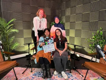 4 women smile at the camera. " sit infront of the other 2, who have their hands on the backs of their chairs, leaning in close. The first women holds a clapperboard reading "Path to Uni: Clearing special"