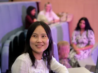An international student looks up from her seat in a lecture hall and smiles at the person approaching