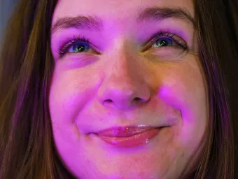 The camera leans close to the face of a student bathed in a warm purple light. She looks up, smiling expectantly