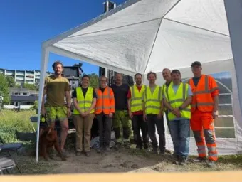 Group of eight people wearing high-visibility vests standing beneath a temporary canopy at an outdoor work site, with equipment and a dog nearby