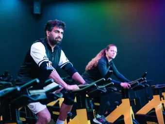 Two people taking part in an indoor spin cycling class on stationary bikes.