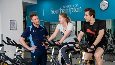 Two students working out on exercise bikes in a gym, assisted by a personal trainer.