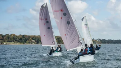 Three small sailing boats, each with a crew of three, on open water beneath blue skies. An area of trees is visible in the background.