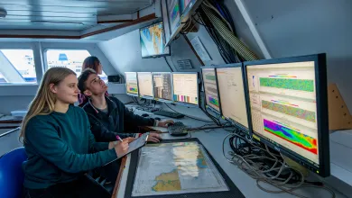 Two students sit at a desk inside a room on a boat. They observe a bank of computer screens displaying various data and information.