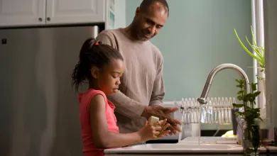 A father smiling down at his young daughter as she washes her hands carefully with soap.