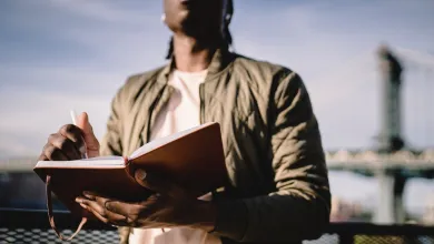 A Black man writes on a notepad as he stands outside, a blue sky and a bridge behind 