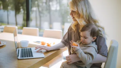 A woman sitting at a table using a laptop with one hand and a child sitting on her knee eating an orange 