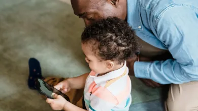 Young child playing on a phone as an adult watches them over their shoulder looks 