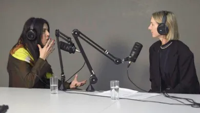 Two people sit behind a desk. Both are wearing large headphones and have large professional microphones in front of them.