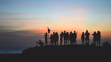 A group of people as silhouettes against a colorful sunset sky, some taking photos and holding a flag, on a rocky lookout.
