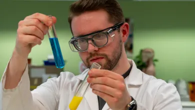 Nicholas in a lab coat and protective eyewear. He's holding two test tubes; one is filled with blue liquid, the other filled with yellow liquid.