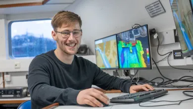 Daniel Hampton is sitting at the work station on Research Vessel Callista. There are screens displaying coulourful charts in the background.