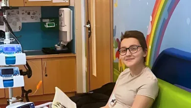 Gio sitting on a colourful bench in a hospital room, reading a book and smiling, with a medical device and snacks nearby.