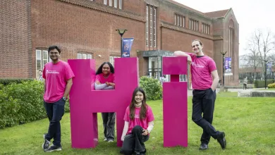 Four students leaning on large pink letters spelling Hi on the grass outside the Hartley library.