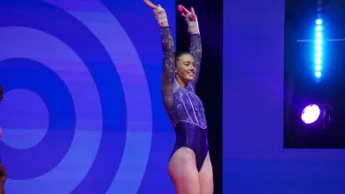 Elite athlete Emily Burke in a purple and silver leotard stands with her arms raised, smiling beneath stage lights after completing her routine.