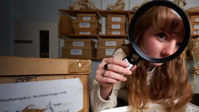 An archaeology student in an archives room, using a magnifying glass to inspect a tray of bones and fossils. The student has red hair and is wearing a cream-coloured knitted sweater. There are more fossils and bones in the background, as well as shelves filled with boxes.