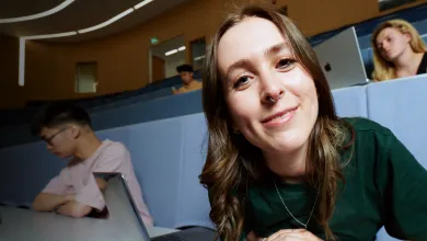 A student smiling while sat in a lecture theatre, with a laptop open on the desk in front of them.