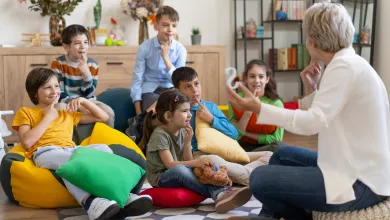 A group of primary school age children are sitting on bean bags on the floor. They are engaged watching a speech therapist talking to them and showing them a large letter ‘S’. They are all touching their chin with an index finger.