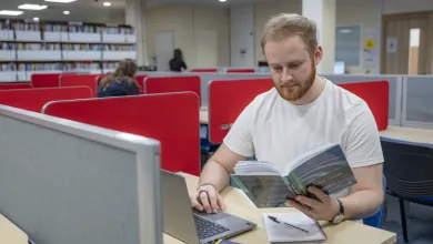 Public Health student, Jack Coles working at a desk in the library. He is reading a book and typing on a laptop keyboard.  