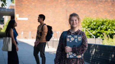 A student smiling while stood outside and holding a closed laptop. There are two more students talking to each other in the background.
