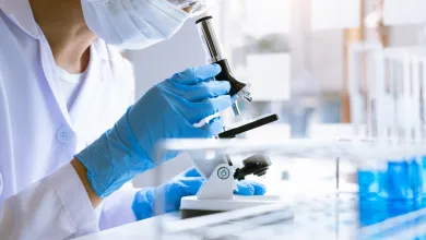 A person in a lab coat using a microscope, wearing a face mask and blue gloves.