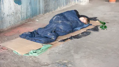 Image shows someone sleeping rough. They're sleeping on the streets in a blue sleeping bag on top of some cardboard.