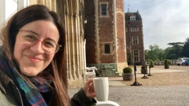 A image of Chiara smiling at the camera. She's sat down outside and is holding a white mug. There's a brick building and grounds behind here.