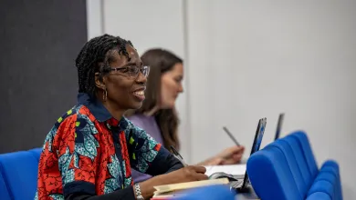 Ogene Adoh sat in a lecture classroom. She has a laptop and notebook in front of her and is looking towards the front of the classroom. 