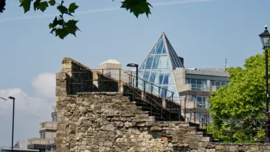 View of a historic stone wall with staircase and metal railings in the foreground, juxtaposed against a modern building featuring a glass pyramid roof. The scene includes leafy tree branches above and a traditional street lamp on the right, highlighting the contrast between medieval and contemporary architecture in an urban setting.
