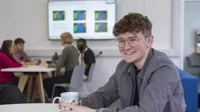 Samuel sitting at a table in a communal area. He has a mug in his hands and is smiling for the camera. 