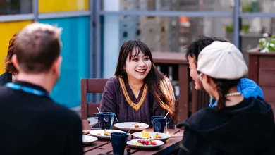 Students sitting outside at a table chatting. There are plates of food and mugs on the table.