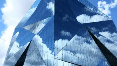 A modern glass building with angular design reflecting the sky and clouds.