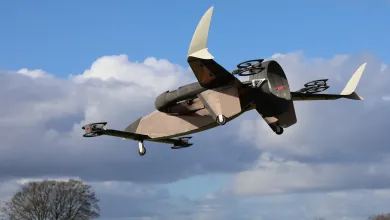 A drone with vertical stabilisers and multiple rotors flying through a blue sky with scattered clouds, above a green field dotted with trees.