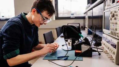 Biomedical engineering PhD student wearing safety goggles soldering a circuit board in an electronics lab.
