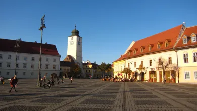 Small, Romanian market square with a clock tower, cobbled paving and painted white buildings, blue sky overhead.
