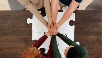 Hands of four people stacked over a white puzzle piece on a wooden table.
