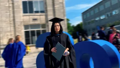 A person wearing graduation robes and a mortarboard stands outdoors holding a certificate folder, with large blue sculpture letters and university buildings in the background.