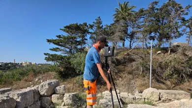 Ben, an archaeology and history BA student, uses surveying equipment on a rocky outcrop surrounded by trees in the mid-day sun